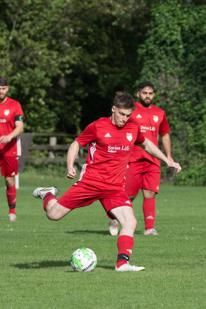 Ein Fußballspieler im roten Trikot beim Schuss auf dem Spielfeld, zwei weitere Mitspieler im Hintergrund