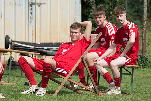 Drei Fußballspieler in roten Trikots sitzen auf Stühlen und entspannen sich während einer Pause.
