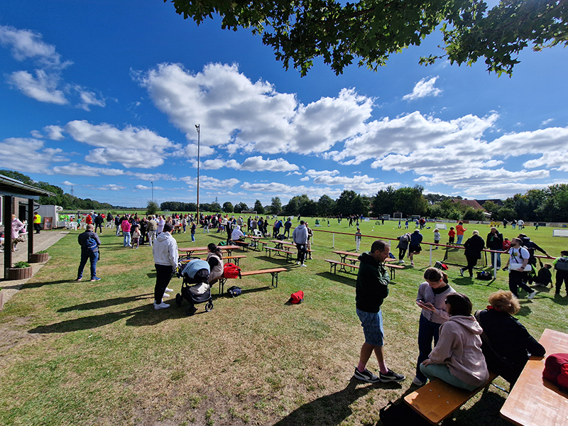 Eine weite Aufnahme eines Fußballplatzes mit vielen Menschen und einigen Tischen. Es sind Kinder und Erwachsene zu sehen, die sich unterhalten oder die Spiele beobachten, während im Hintergrund auf dem Spielfeld Fußball gespielt wird. Der Himmel ist blau mit einigen Wolken.